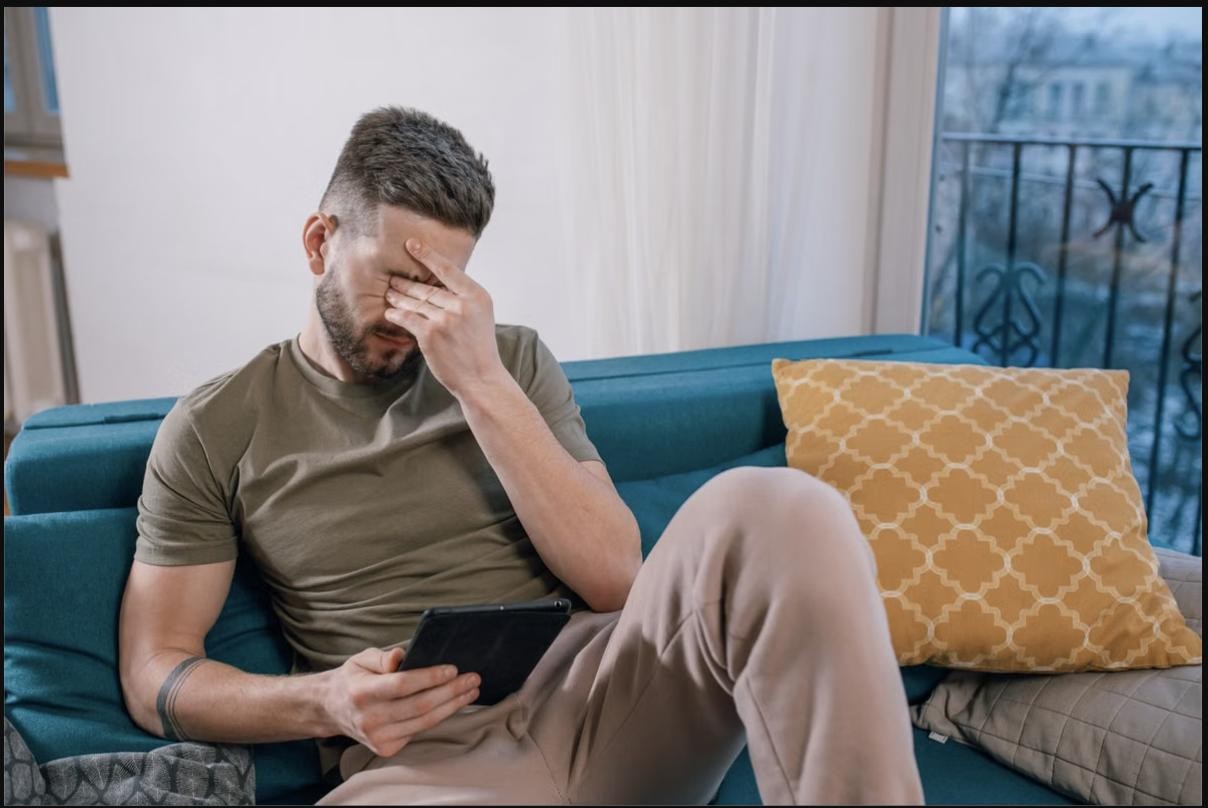 A father sitting on the couch, overwhelmed, holding his head while looking at a tablet