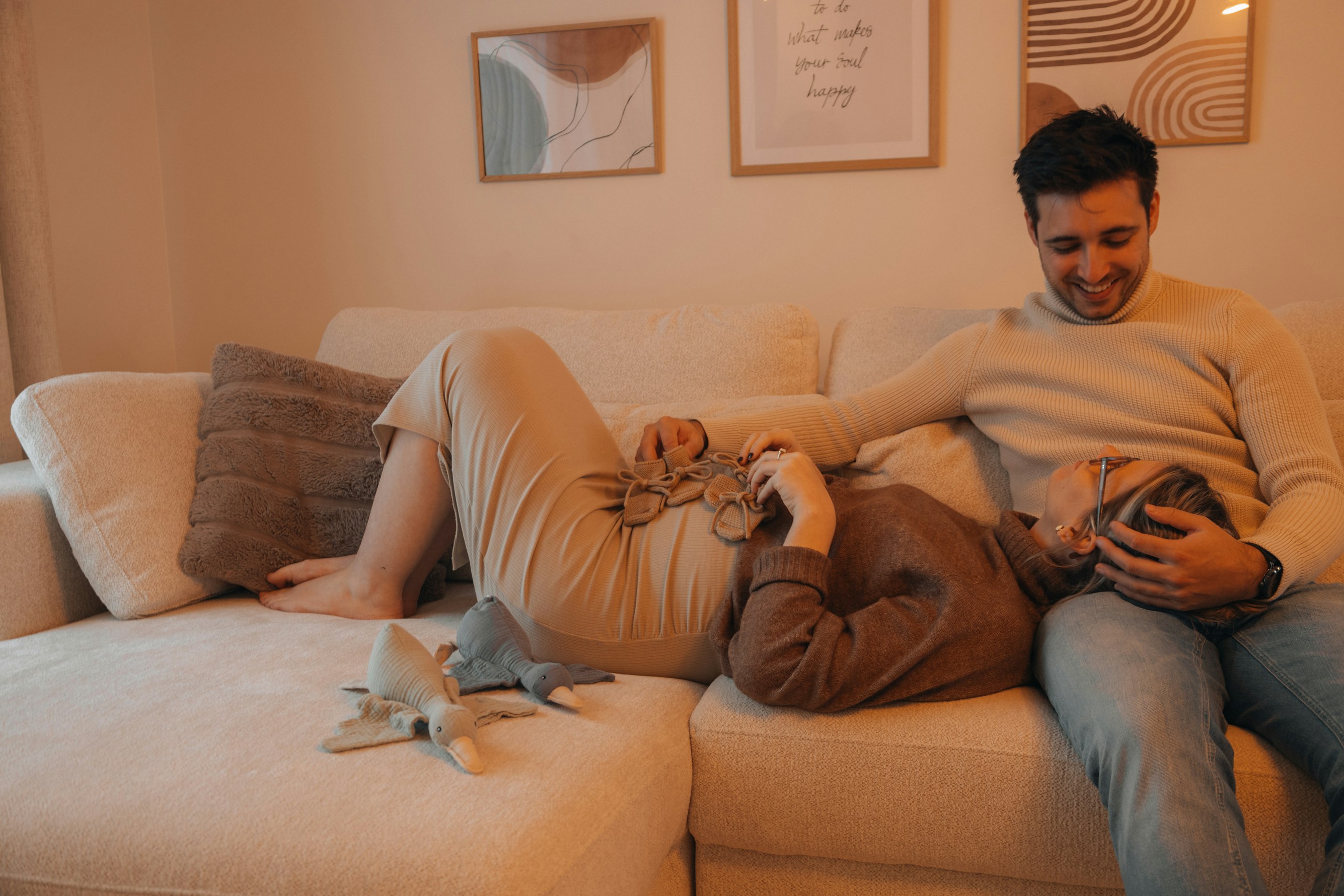 An expecting couple sitting close together, the partner resting a hand on her pregnant belly in soft natural light